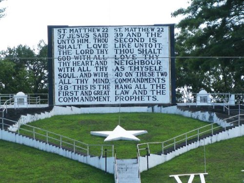 A large sign displaying biblical verses about loving God and neighbors, set on a grassy hill with a star shape below.