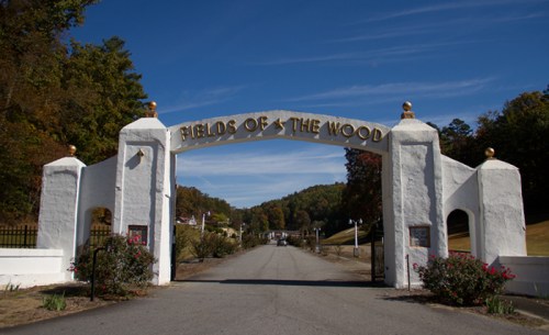 White archway with "FIELDS OF THE WOOD" sign, leading to a scenic road surrounded by trees and hills.