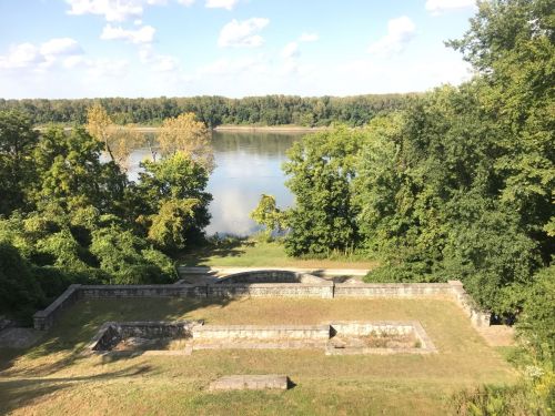 A serene view of a river surrounded by trees, with a stone structure in the foreground and a clear blue sky above.