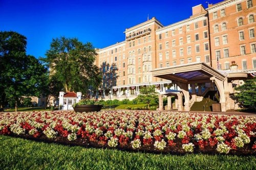 Historic hotel with a grand facade, surrounded by vibrant flower beds and lush green lawns under a clear blue sky.