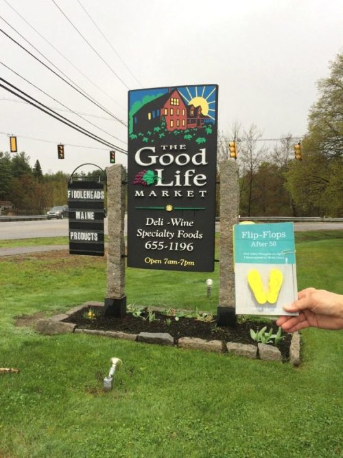 A person holds a yellow flip-flop card in front of a market sign that lists deli, wine, and specialty foods.