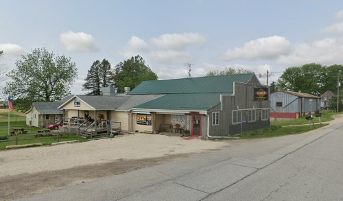 A small rural store with a green roof, surrounded by trees and a gravel road, featuring outdoor seating and a sign.