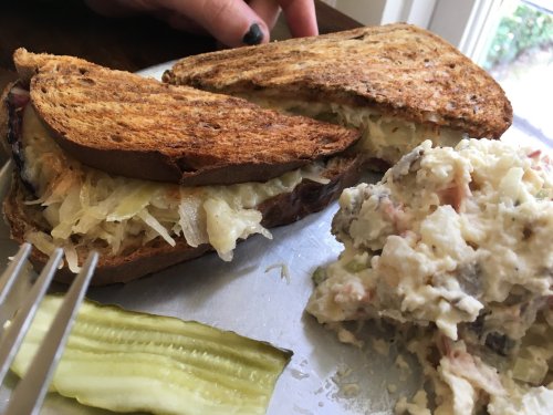 A toasted sandwich filled with sauerkraut, served with a side of potato salad and a pickle slice on a plate.