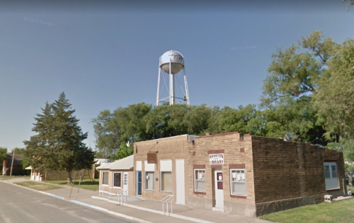 A brick building with a library sign, next to a water tower and surrounded by trees on a quiet street.
