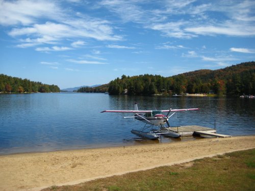 A seaplane is docked on a calm lake surrounded by trees and mountains under a blue sky with scattered clouds.