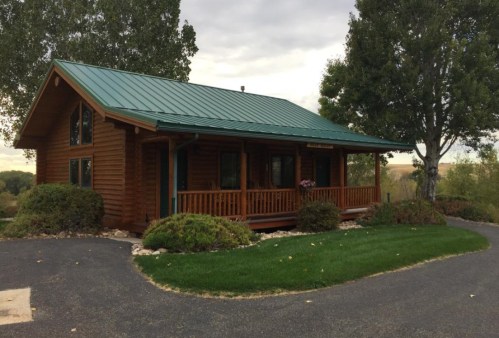 A cozy wooden cabin with a green metal roof, surrounded by trees and a well-maintained lawn.