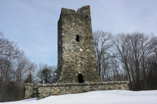 A tall, weathered stone tower stands in a snowy landscape, surrounded by bare trees under a cloudy sky.