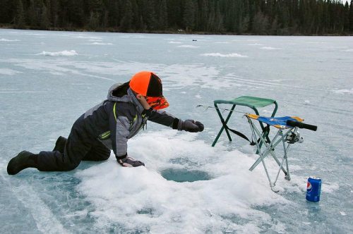 A child in winter gear kneels on an icy lake, reaching into a hole while a chair and a soda can sit nearby.