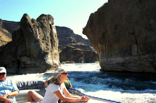 A boat navigating through rocky cliffs on a river, with two people enjoying the scenery under a clear blue sky.