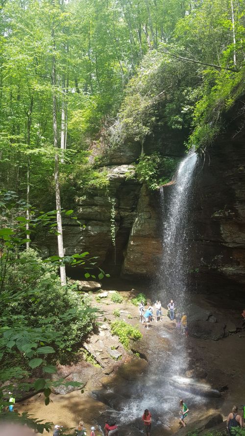 A scenic waterfall cascades down rocky cliffs, surrounded by lush green trees and people enjoying the natural beauty below.