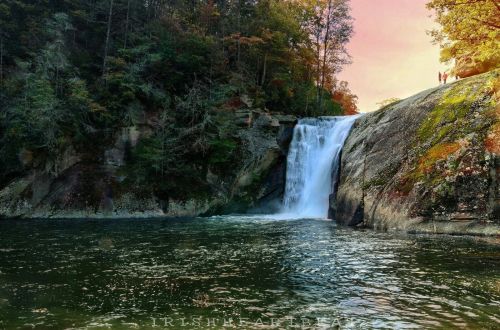 A serene waterfall cascades over rocks into a calm pool, surrounded by autumn foliage and a colorful sky.