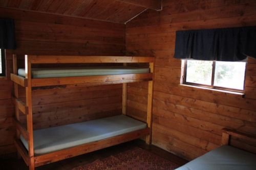 A rustic wooden cabin interior featuring a bunk bed and a single bed, with two windows and wooden walls.