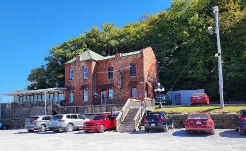 Historic brick building with a green roof, surrounded by trees and parked cars in a gravel lot. Clear blue sky above.