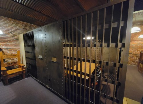 A dimly lit room featuring a large, barred metal door and a wooden table surrounded by chairs. Brick walls are visible.