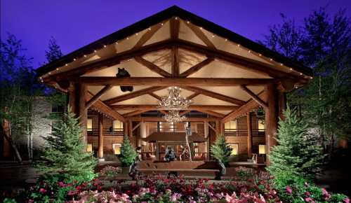 A beautifully lit lodge entrance with wooden beams, surrounded by trees and colorful flowers at dusk.