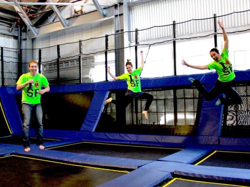 Three people in bright green shirts jump on trampolines in a lively indoor trampoline park.