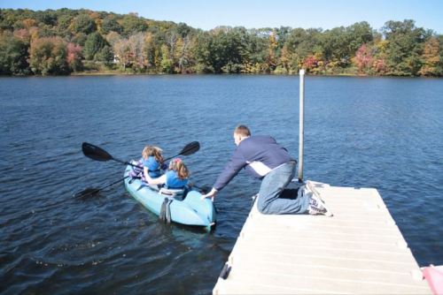 A man helps two children in a kayak as they prepare to paddle on a calm lake surrounded by autumn trees.