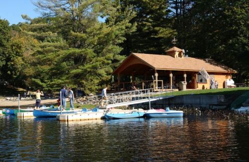 A lakeside scene with a wooden pavilion, people by the dock, and blue kayaks on calm water surrounded by trees.