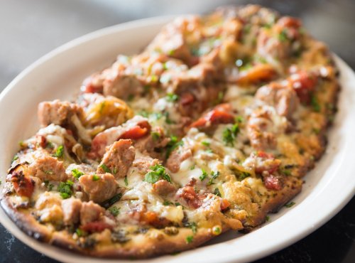 A delicious flatbread pizza topped with sausage, tomatoes, cheese, and fresh herbs on a white plate.