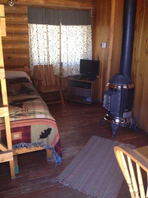 Cozy cabin interior with wooden furniture, a bed, a TV, and a wood stove, featuring warm lighting and rustic decor.