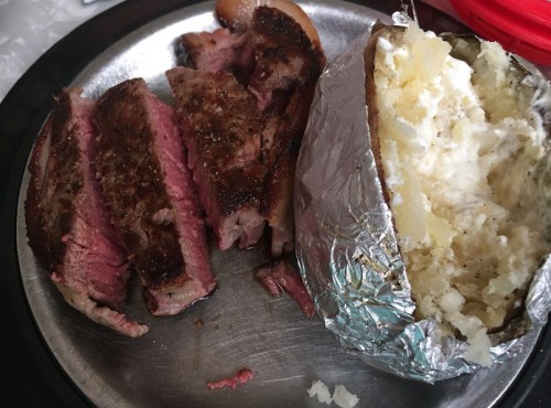 A plate with sliced steak and a baked potato wrapped in foil, showcasing juicy pink meat and creamy filling.