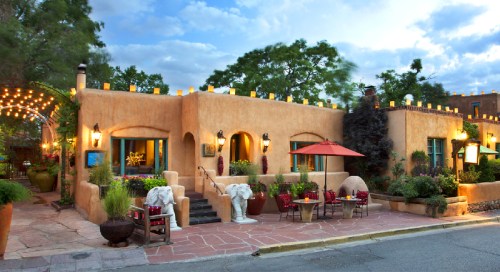 A charming adobe-style restaurant with outdoor seating, surrounded by greenery and decorative lights under a cloudy sky.