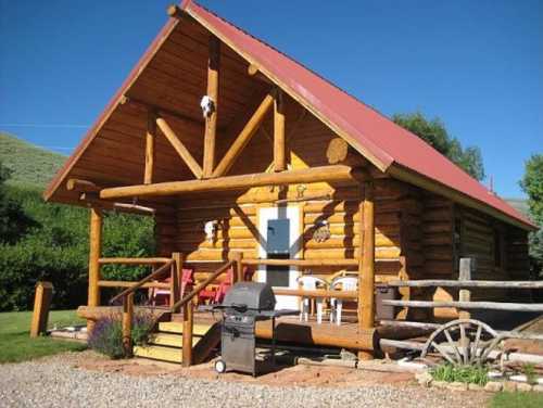 A rustic log cabin with a red roof, surrounded by greenery and featuring a porch and outdoor grill.