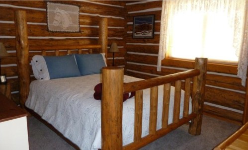 Cozy log cabin bedroom featuring a wooden bed, blue pillows, and warm lighting from a window.