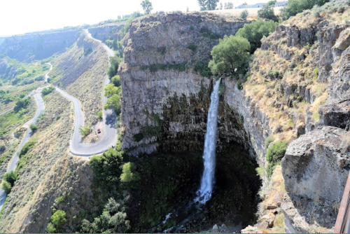 A waterfall cascades down a rocky cliff, surrounded by greenery and a winding road in the background.