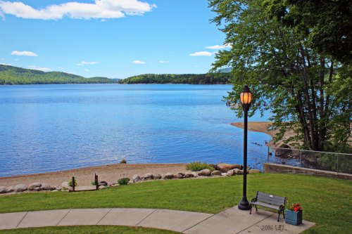 A serene lake view with clear blue water, green trees, and a sandy shore under a bright sky. A bench and lamp are in the foreground.