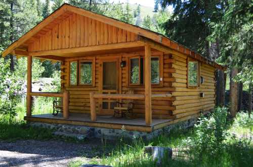 A cozy wooden cabin surrounded by greenery, featuring a porch with chairs and large windows.