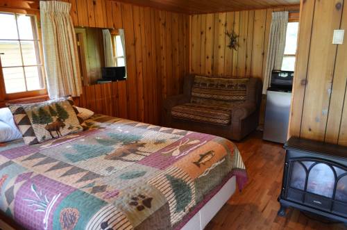 Cozy cabin interior featuring a bed with a patterned quilt, a sofa, and wooden walls. A small fridge is visible.