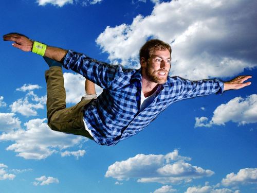 A man in a blue checkered shirt and khaki pants is flying through a bright blue sky with clouds.