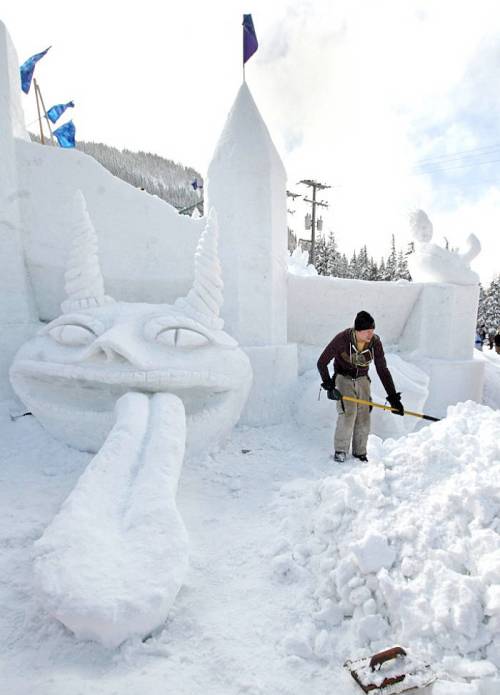 A person shovels snow near a large, whimsical snow sculpture with a face and horns, set against a snowy landscape.