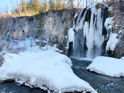 A frozen waterfall surrounded by snow-covered rocks and trees, with sunlight illuminating the scene.