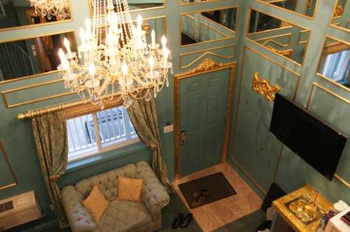 A stylish room with a chandelier, green walls, gold accents, a plush sofa, and a TV, viewed from above.