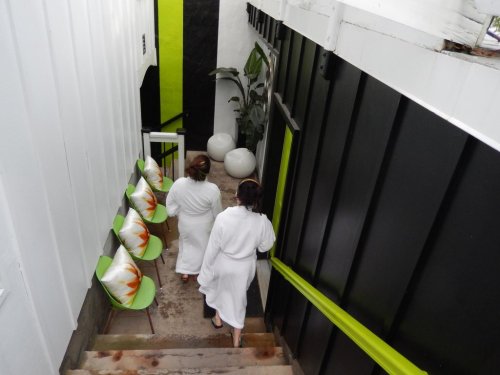 Two women in white robes walk down a staircase, flanked by green chairs and potted plants in a modern interior.