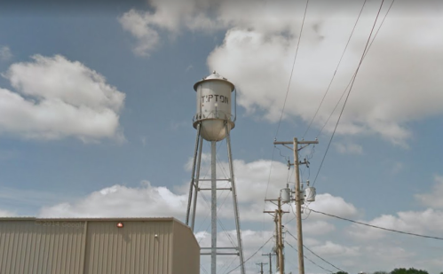 A tall water tower stands beside a building, with power lines and a cloudy sky in the background.