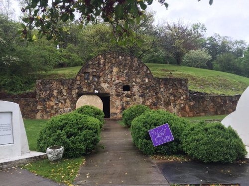 A stone structure with a rounded arch entrance, surrounded by green bushes and trees, under a cloudy sky.