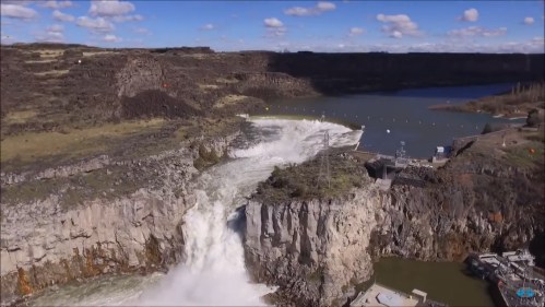 Aerial view of a waterfall cascading over rocky cliffs into a river, surrounded by green hills and blue sky.