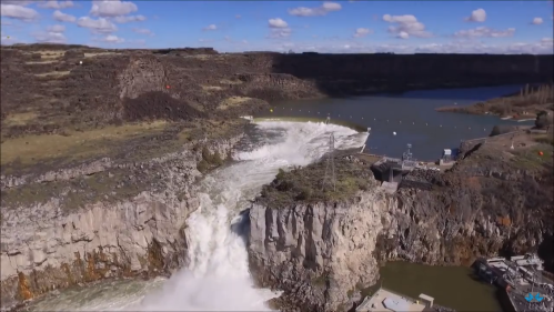 Aerial view of a waterfall cascading over rocky cliffs into a river, surrounded by green hills and blue sky.