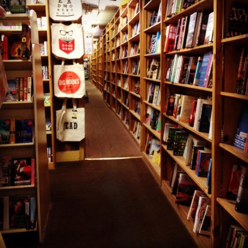 A cozy bookstore aisle lined with wooden shelves filled with various books and tote bags hanging on the side.