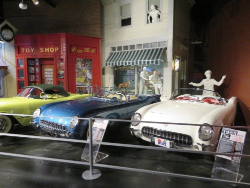 Three classic cars on display in a museum, with a toy shop and a house in the background.
