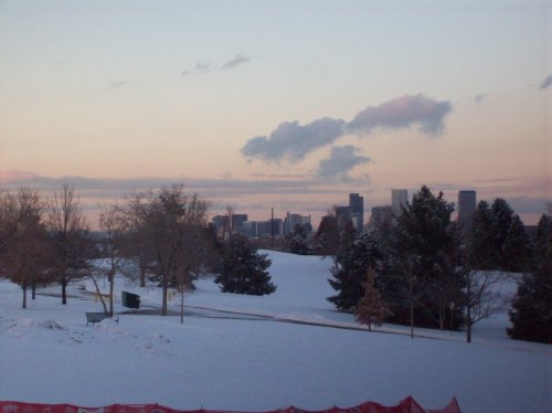 A snowy landscape with trees in the foreground and a city skyline in the background during sunset.