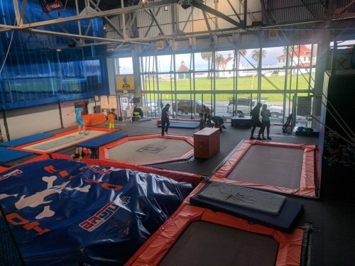 A spacious indoor trampoline park with various trampolines and people jumping and playing. Large windows show a view outside.