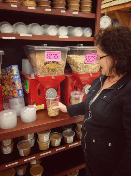 A person fills a jar with granola from a red dispenser in a store, surrounded by various containers of snacks.