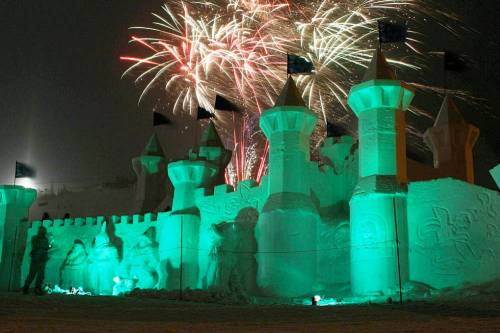 A snow castle illuminated in green light, with fireworks bursting in the night sky above it.