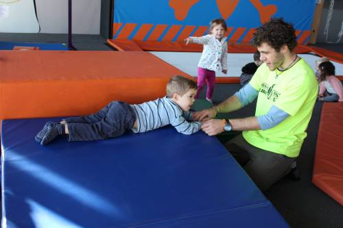 A child lies on a mat while an adult assists him; another child plays in the background at a gym.