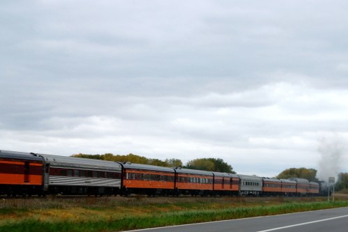 A vintage train with orange and silver cars travels along a track beside a grassy field under a cloudy sky.