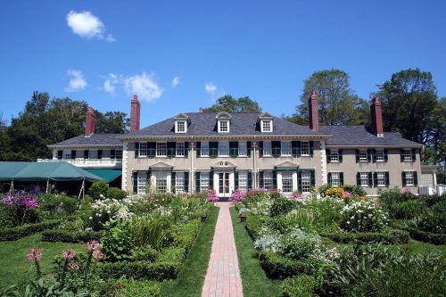 A large, elegant mansion surrounded by lush gardens and colorful flowers under a clear blue sky.